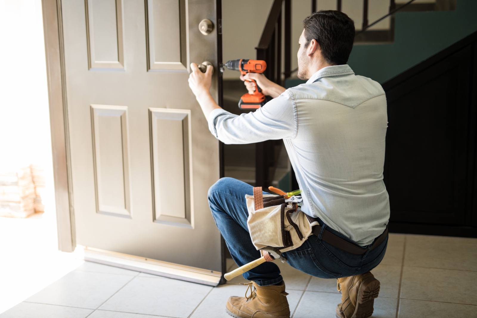 Handyman in light blue shirt using cordless drill on door while crouching with tool belt