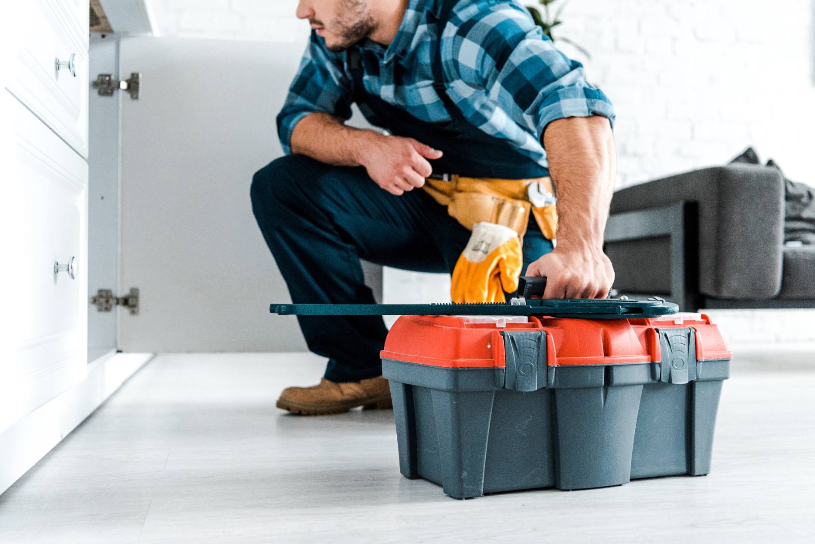Plumber in blue plaid shirt opening red and gray toolbox while crouching in kitchen
