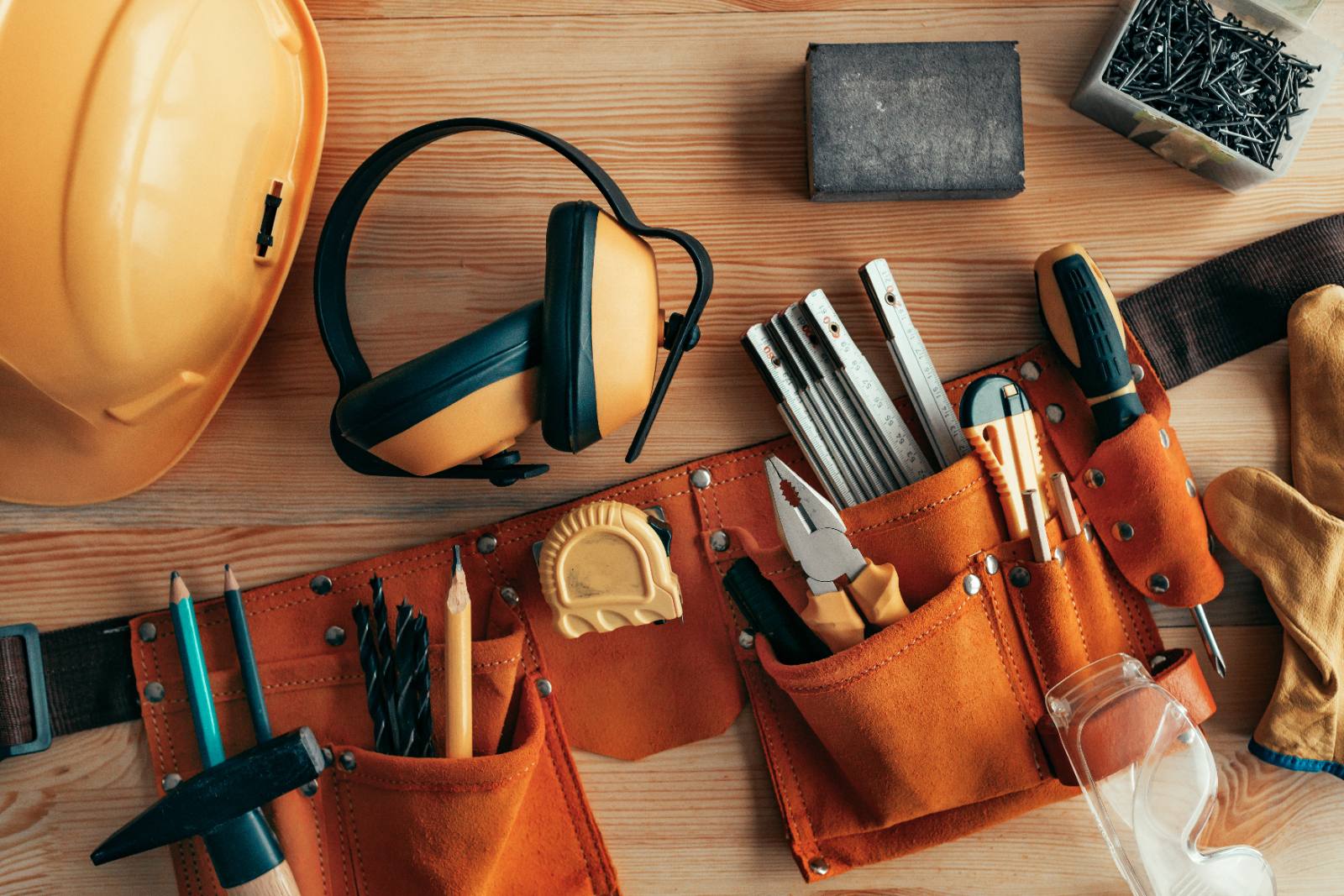 Construction tools and safety equipment arranged on wooden surface including hard hat, headphones, tool belt, and hardware